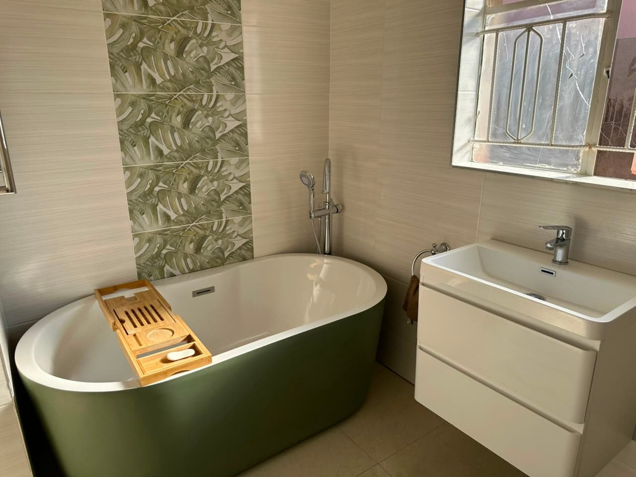 Olive green freestanding bath with a floor-mounted mixer, set against a monstera-leaf feature tile panel beside a wall-hung vanity