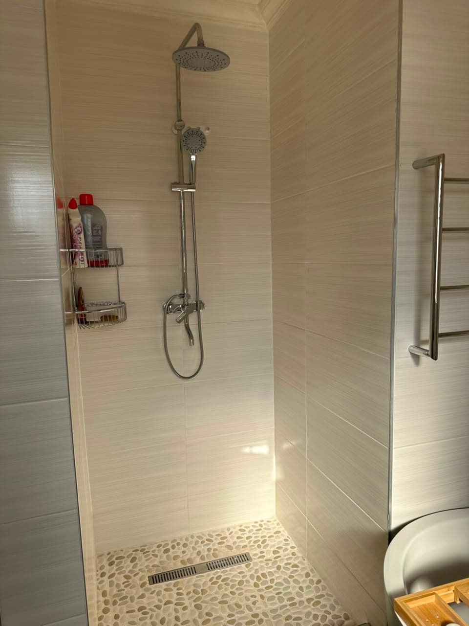Cream tiled shower with a rain head and handheld riser, a pebble mosaic floor and a stainless linear drain