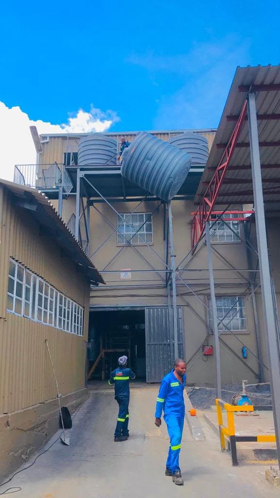 Water storage tank being hoisted onto an elevated steel stand above the yard of an industrial building
